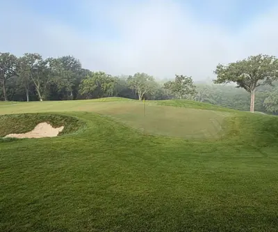 Fog over the treeline behind a green with yellow flag and greenside bunker at Quarry Oaks Golf Club in Ashland, Nebraska
