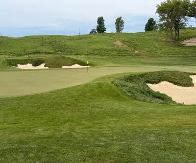 Green with a yellow flag guarded by deep sand bunkers below grassy hills at Quarry Oaks Golf Club in Ashland, Nebraska