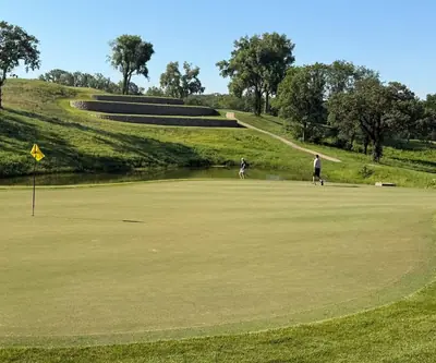 Yellow-flag green with golfers by a pond and terraced stone walls beyond at Quarry Oaks Golf Club in Ashland, Nebraska