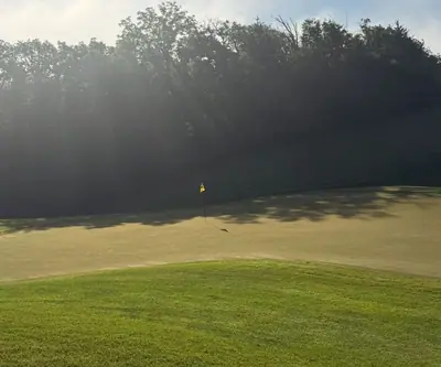 Sunbeams filtering through trees onto a green with a yellow flag at Quarry Oaks Golf Club in Ashland, Nebraska