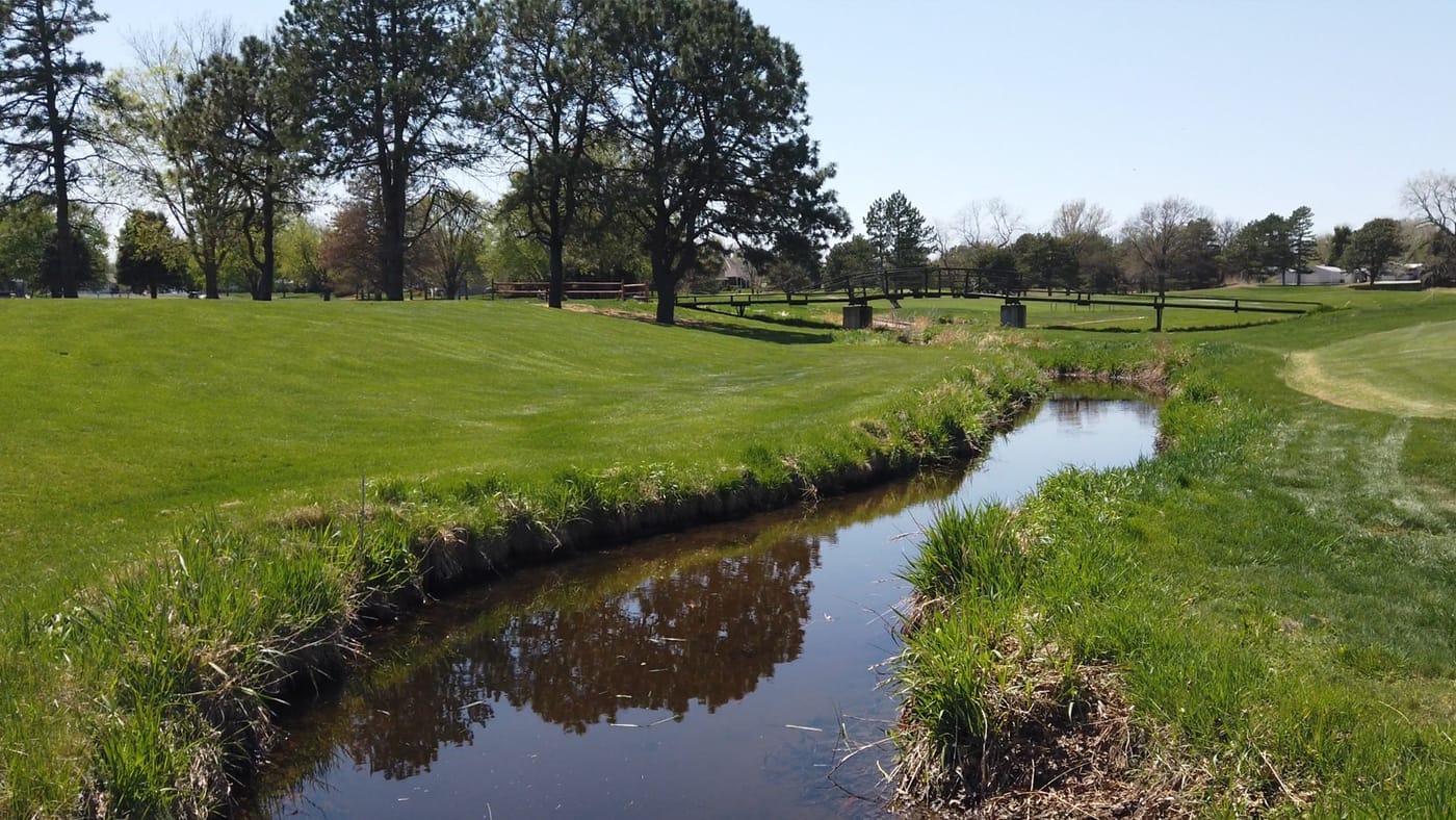 Creek winding through fairways toward a wooden footbridge at Poco Creek Golf Course in Aurora, Nebraska