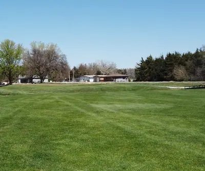 Wide fairway with a black-railed footbridge and low buildings beyond at Poco Creek Golf Course in Aurora, Nebraska