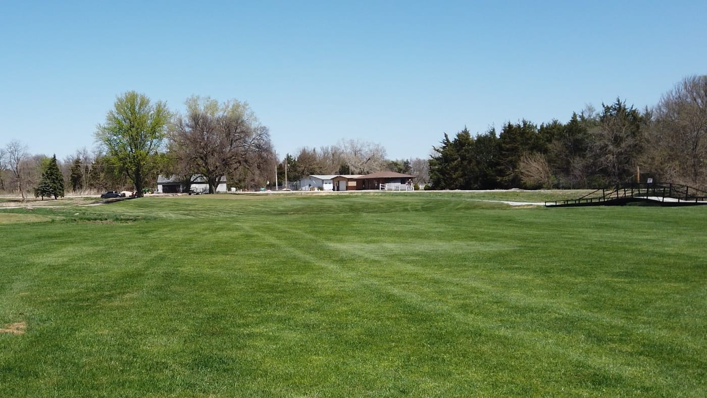 Wide fairway with a black-railed footbridge and low buildings beyond at Poco Creek Golf Course in Aurora, Nebraska