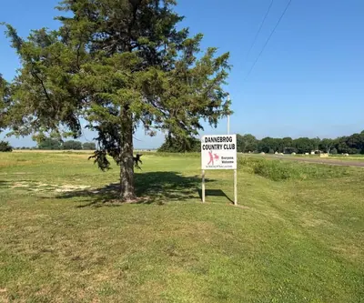 Dannebrog Country Club 'Everyone Welcome' sign beside an evergreen tree along a country road in Dannebrog, Nebraska