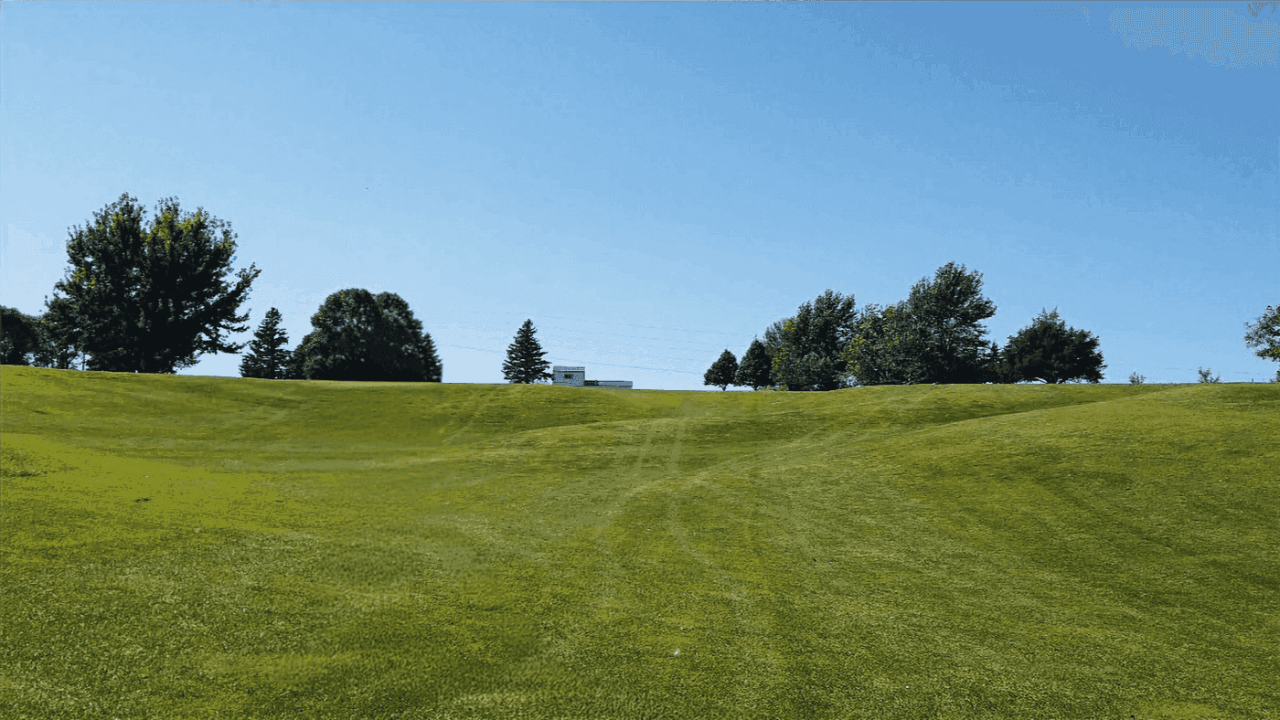 Green grassy field with rolling hills, scattered trees in the background, and a small white building under a clear blue sky. Calm and serene.  Hole #3 at Gibbon Valley View Golf Course in Gibbon, Nebraska