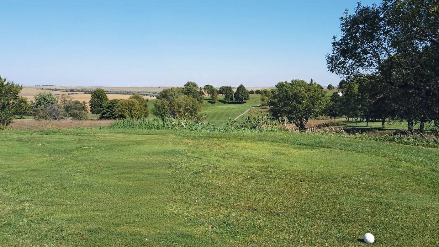 A golf ball on a green fairway, surrounded by trees and open fields under a clear blue sky. Calm and serene landscape. Hole #5 at Gibbon Valley View Golf Course in Gibbon, Nebraska