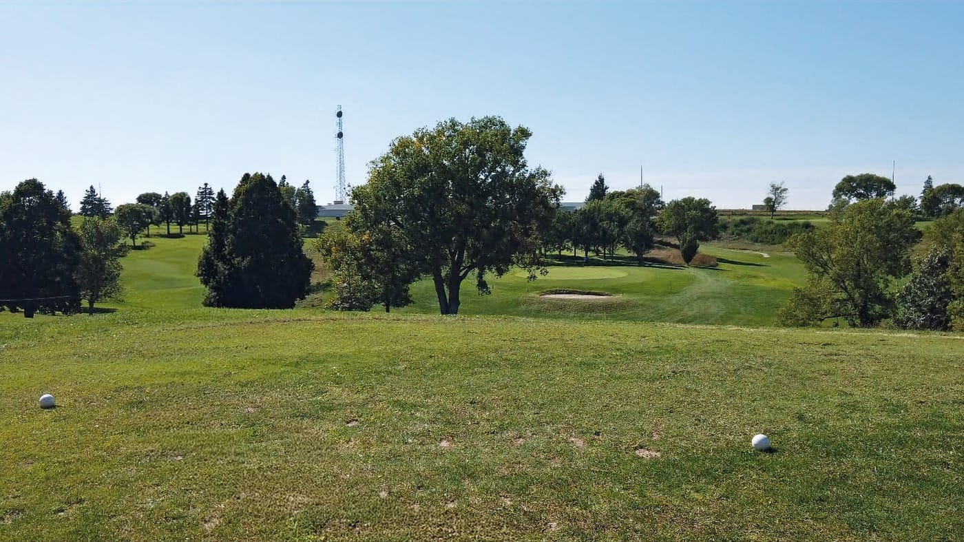 Green golf course with trees and a sand trap in the background. Two golf balls rest on the grass under a clear blue sky. Peaceful setting. Hole #6 at Gibbon Valley View Golf Course in Gibbon, Nebraska
