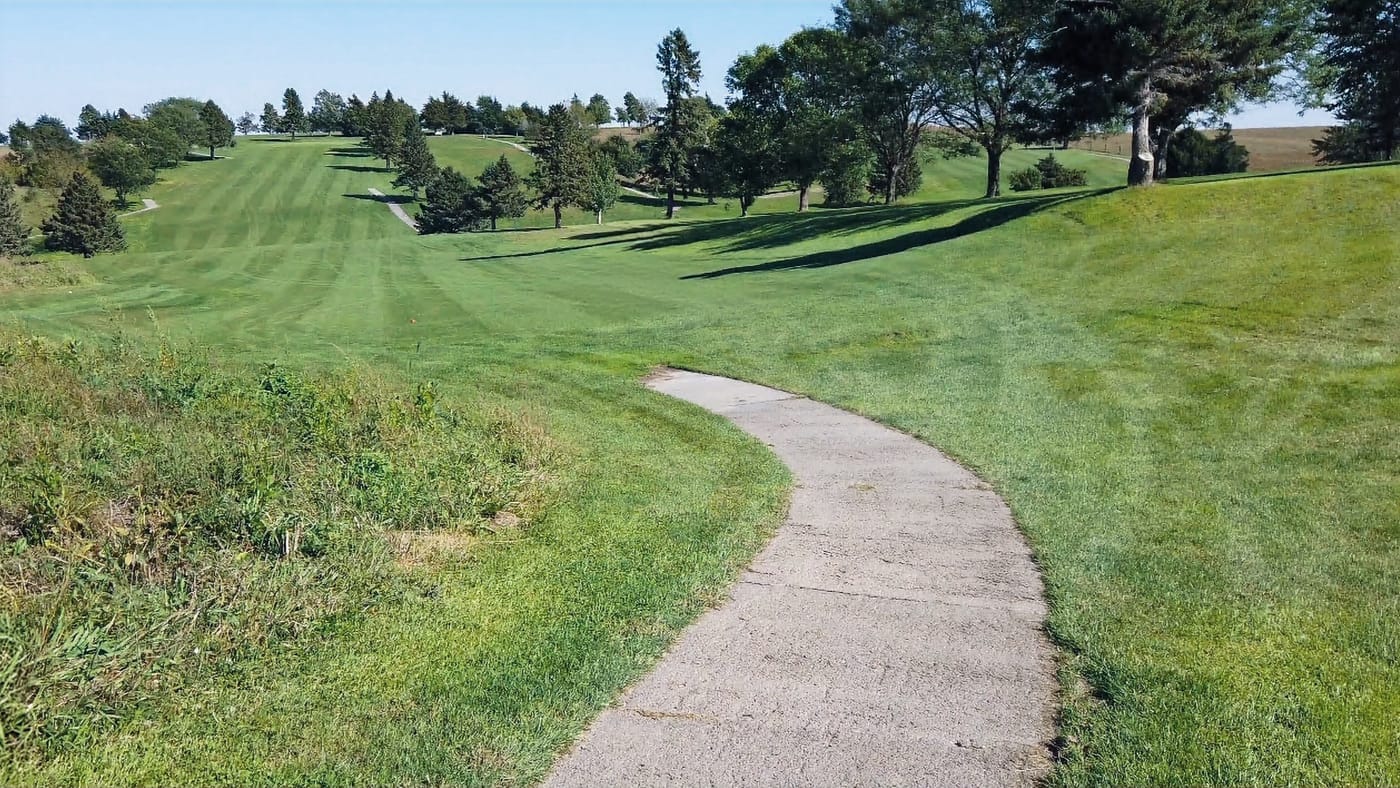 A winding concrete path cuts through a lush, green golf course with neatly trimmed grass and scattered trees under a clear blue sky.  Hole #8 at Gibbon Valley View Golf Course