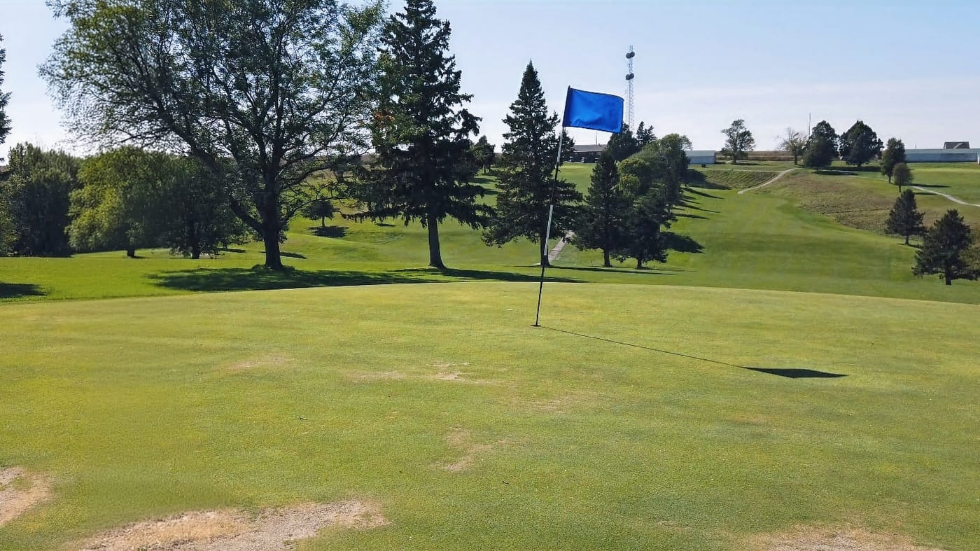 Golf course with green grass and blue flag on a pole, surrounded by trees under a clear blue sky. Relaxing and serene atmosphere.  Hole #6 at Gibbon Valley View Golf Course