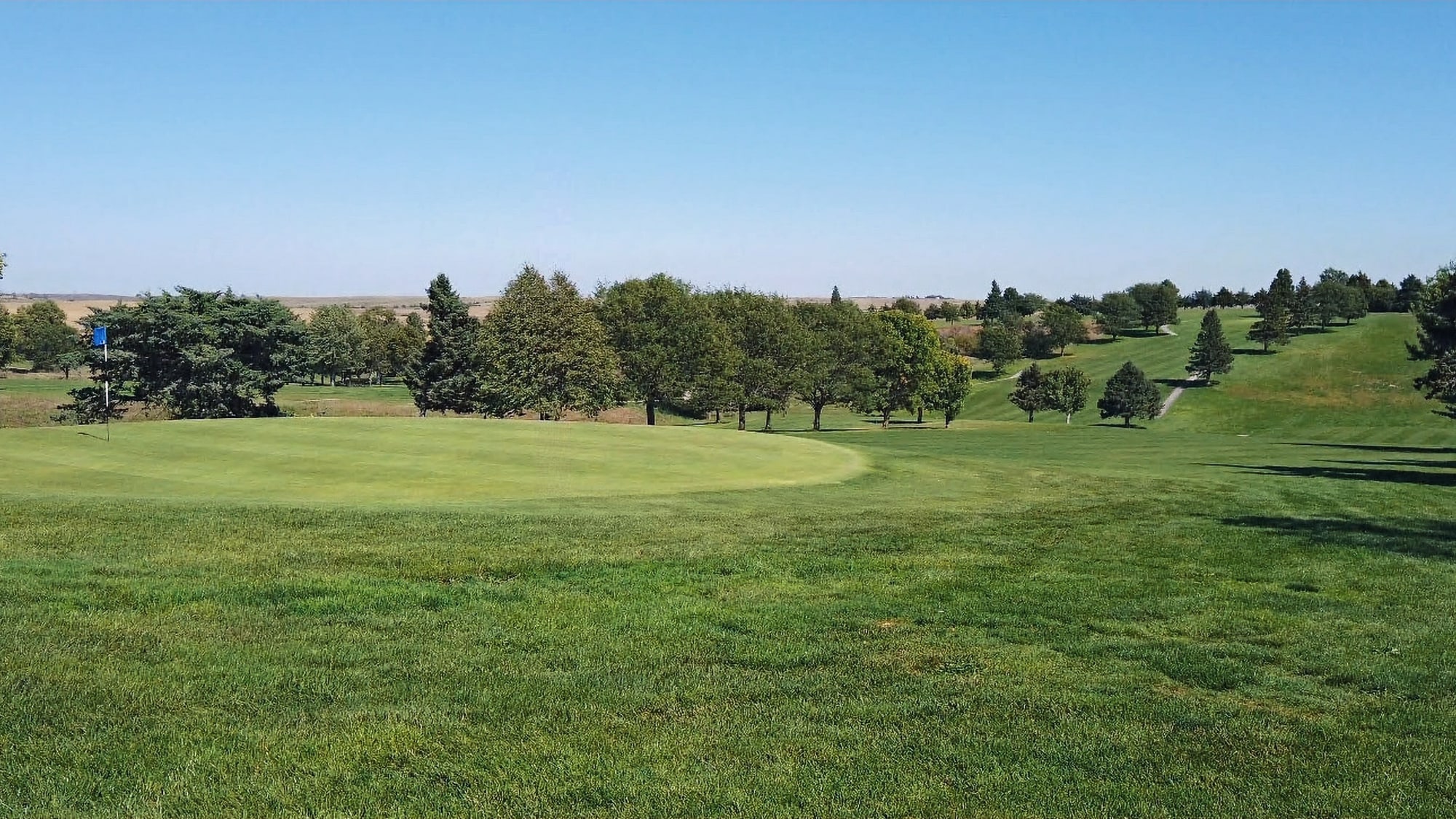 Golf course with a lush green fairway and putting green. Trees line the background under a clear blue sky, creating a serene scene.  Hole #9 at Gibbon Valley View Golf Course