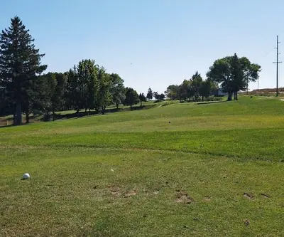 Golf course scene with green grass, scattered trees, and a white golf ball on the ground. Blue sky above, creating a serene atmosphere.  Hole #4 at Gibbon Valley View Golf Course in Gibbon, Nebraska