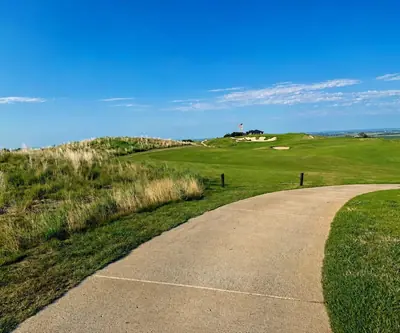 Cart path curving toward a fairway, bunkers, and a hilltop American flag at Landmand Golf Club in Homer, Nebraska