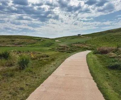 Concrete cart path through native grasses toward bunkers and a hilltop shelter at Landmand Golf Club in Homer, Nebraska