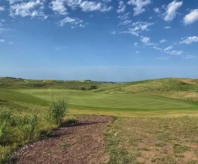 Dirt path leading to a large rolling green with a yellow flag amid grassy hills at Landmand Golf Club in Homer, Nebraska