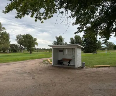 Small white shelter with a bench and trash cans beside a gravel lot at Loup City Golf Club in Loup City, Nebraska