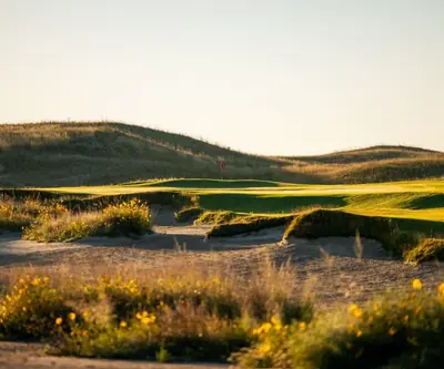 Red flag on a green beyond sandy bunkers and yellow wildflowers in golden light at GrayBull Golf Club in Maxwell, Nebraska