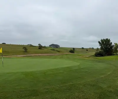 Green with yellow flag and rolling grassy hills under an overcast sky at The Greens At Prairie Hills in Pleasanton, Nebraska