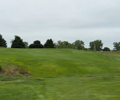 Uphill approach to a green with yellow flag framed by evergreens at The Greens At Prairie Hills in Pleasanton, Nebraska