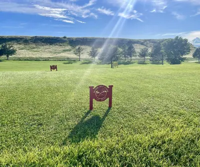 Maroon tee sign with course logo on a sunlit tee box below a grassy bluff at Hillside Golf Course in Sidney, Nebraska