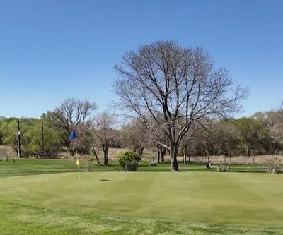 Putting green with a blue flag, bare spring trees and a small shelter at St. Paul Country Club in St. Paul, Nebraska