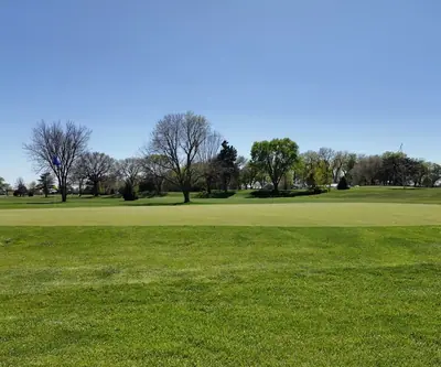 Wide putting green with blue flag and spring trees under clear sky at St. Paul Country Club in St. Paul, Nebraska